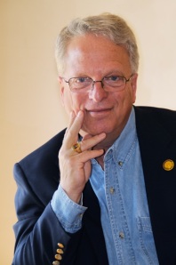 Light-skinned man with short blond hair receding at the temple. Wears glasses, a pale blue oxford shirt and navy blue jacket with gold-colored buttons at the cuff. 